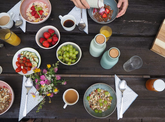 A breakfast table with bowls of cereal, strawberries, grapes, sliced vegetables, drinks, cups, cutlery, napkins, and a small vase of flowers. A hand pours milk into a bowl.