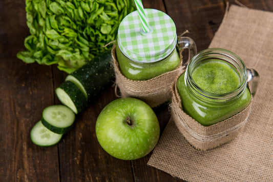 Deux pots Mason de smoothie vert sur de la toile de jute, avec de la pomme verte, des tranches de concombre et de la laitue sur une table en bois.