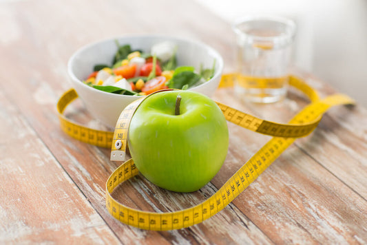 Une pomme verte, un mètre ruban jaune, un bol de salade et un verre d'eau sont posés sur une table en bois.