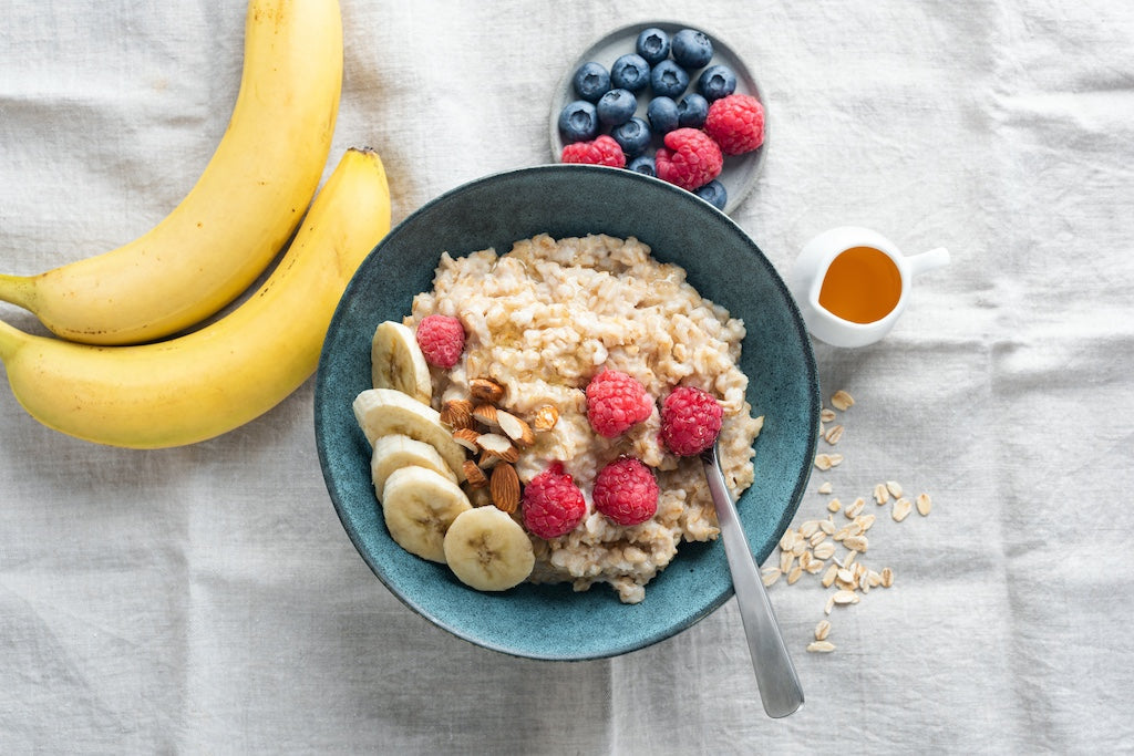 A bowl of porridge with banana slices, raspberries, and nuts, next to it two bananas, a plate of blueberries and raspberries, a small jug of honey, and scattered oat flakes on a cloth.