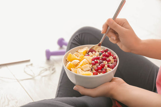 Une personne tient un bol de flocons d'avoine avec des tranches d'orange, de la banane et des baies rouges - et savoure ce petit-déjeuner réconfortant assis sur le sol à côté d'haltères violets, parfaits pour ceux qui veulent perdre du poids.