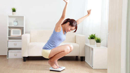 Dans un salon lumineux avec un canapé blanc et des plantes vertes, une femme est assise sur une balance de salle de bain et tend les bras vers le haut avec joie.