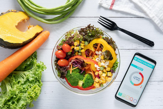 Un bol de salade mélangée et de légumes à côté d'un smartphone avec un compteur de calories quotidien, posé sur une table en bois blanc avec des fruits et légumes frais autour.