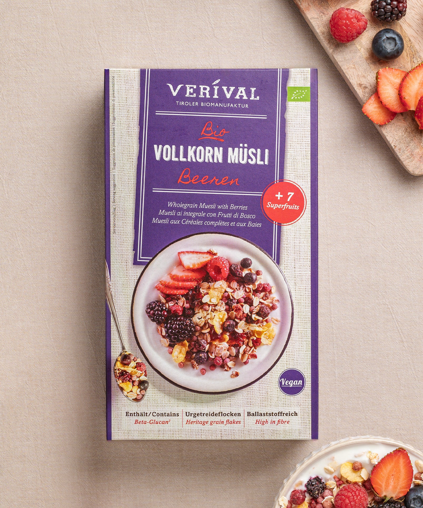 Eine Packung ballaststoffreiches Beeren Vollkorn Müsli von Verival Bio ist neben einer Schüssel mit Müsli und Superfrüchten wie Erdbeeren und Heidelbeeren vor hellem Hintergrund zu sehen.