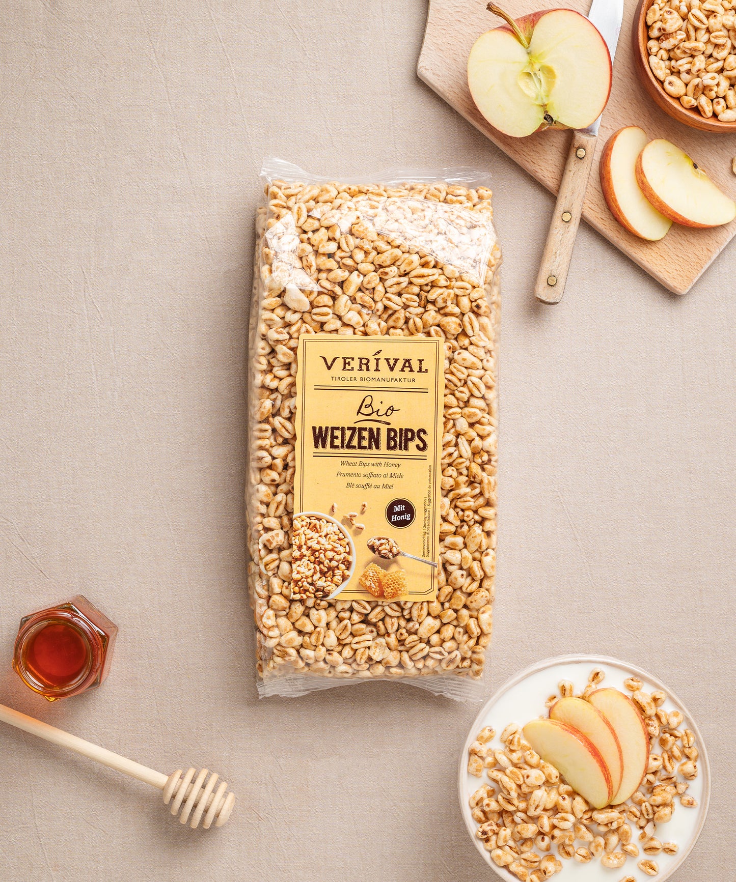 Eine Packung Weizenbips mit Honig von Verival Bio liegt zwischen Apfelscheiben, einer kleinen Schüssel gepufftem Weizenmüsli, einem Glas Bienenhonig und einem Honiglöffel.