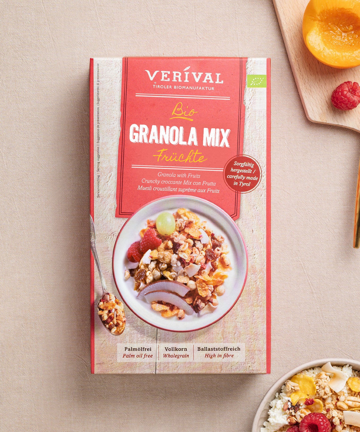 Eine Schachtel Granola Mix Früchte von Verival Bio steht auf einem beigen Untergrund. Darin befindet sich eine farbenfrohe Schale mit Granola, Nüssen und Trockenfrüchten. Daneben steht eine weiße Schale mit proteinreichem Granola, umgeben von verstreuten frischen Früchten.