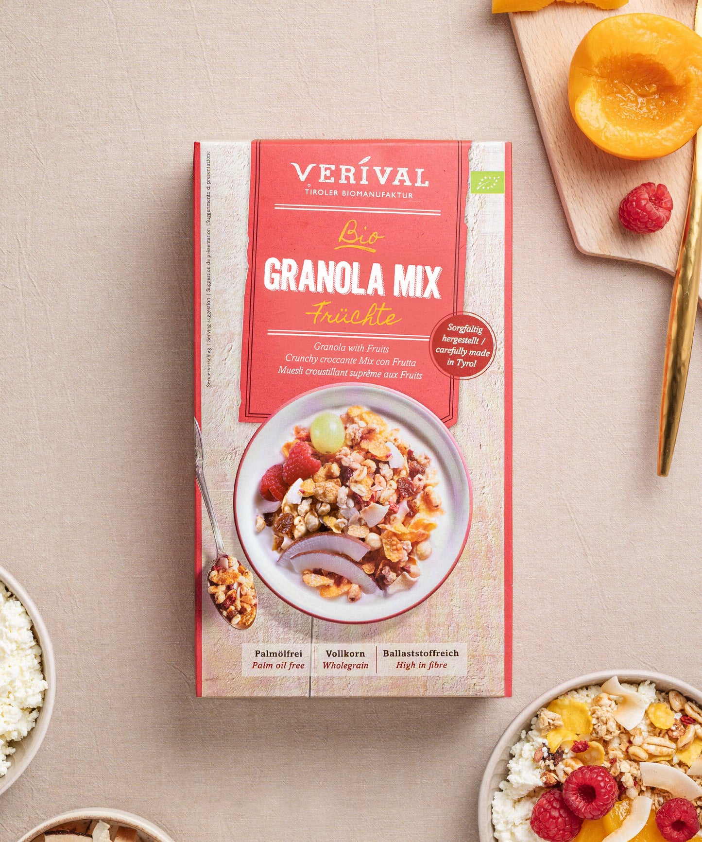 Auf einem Tisch steht eine Packung Granola Mix Früchte von Verival Bio, daneben eine Schüssel mit dem fruchtigen und ballaststoffreichem Granola. Daneben liegen Pfirsichscheiben, Himbeeren und ein Holzlöffel und schaffen damit eine einladende Frühstücksszenerie.
