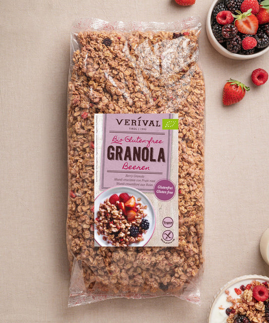 Eine große Tüte Verival Bio GmbH Bio Beeren Granola glutenfrei ruht auf einer beigen Fläche, umgeben von Schalen mit Beeren, Joghurt und Granola.