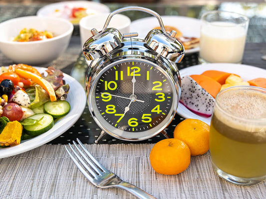 Ein silberner analoger Wecker steht zwischen verschiedenen Frühstücksspeisen, darunter Salat, Obst, Saft und ein Glas Milch auf einem Tisch.