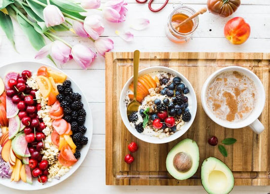 Eine Frühstücksszene mit einer Obst- und Müslischale, einem Teller mit verschiedenen frischen Früchten, einer Avocado, einer Tasse Kaffee, Honig, Tulpen und einem Pfirsich auf einer Holzoberfläche.