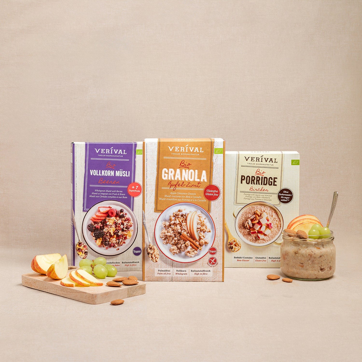 Drei Boxen mit Verival Frühstücksprodukten: Vollkornmüsli, Granola und Porridge, präsentiert mit frischem Obst und Nüssen auf beigem Hintergrund.