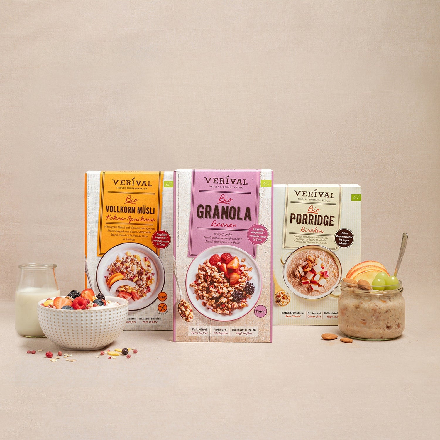 Drei Müsli-Boxen von Verival: Vollkorn Müsli, Granola Berries und Porridge. Abgebildet mit einer Schüssel Müsli, einer Schüssel Porridge, einem Glas Milch und Obst auf beigem Hintergrund.