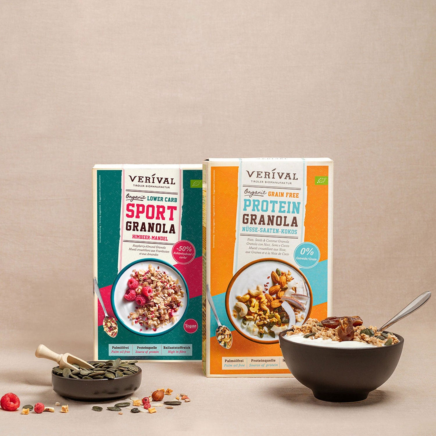 Zwei Schachteln Verival-Granola (Sport Granola und Protein Granola) stehen hinter einer Schüssel Granola mit einem Löffel und einer kleinen Schale mit Körnern vor einem neutralen Hintergrund.