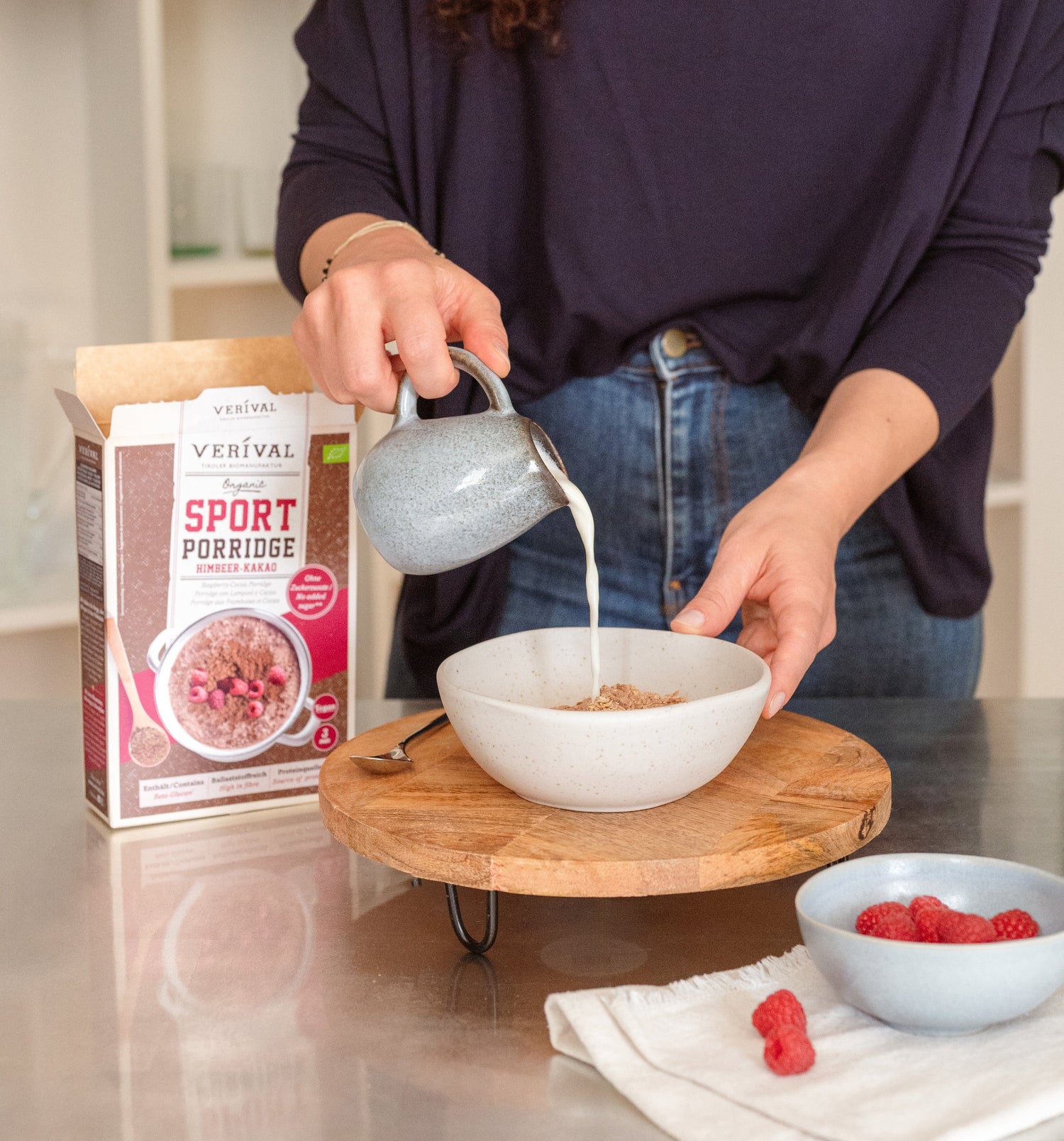 Eine Person gießt Milch in eine Schüssel Brei neben einer Schachtel mit der Aufschrift Sport Porridge und einer kleinen Schüssel mit Himbeeren auf einem Holztablett.