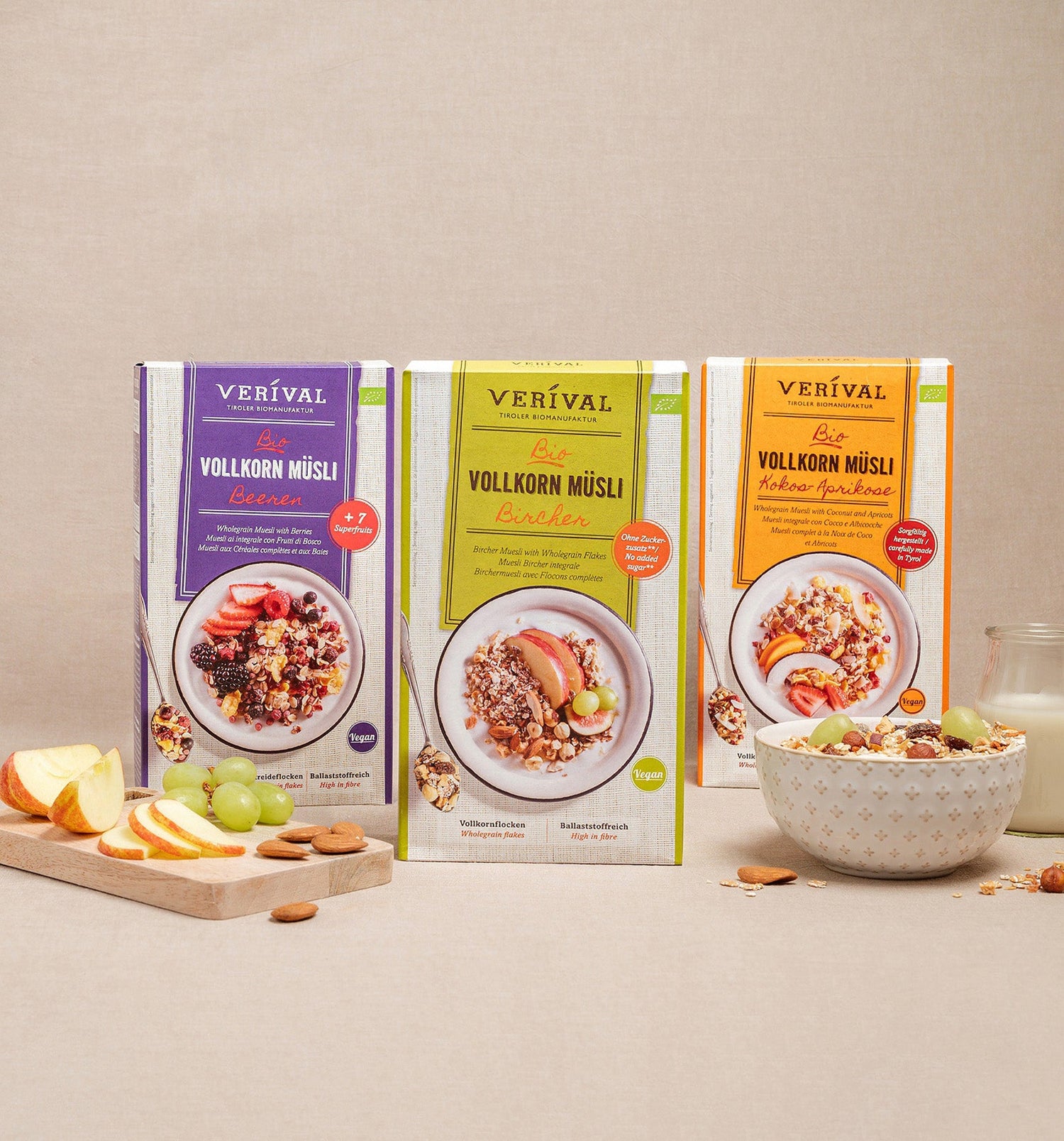 Drei Schachteln Verival Vollkorn Müsli in verschiedenen Geschmacksrichtungen stehen aufrecht auf einer beigen Fläche, daneben eine Schale mit Müsli, Obst und ein Glas Milch.