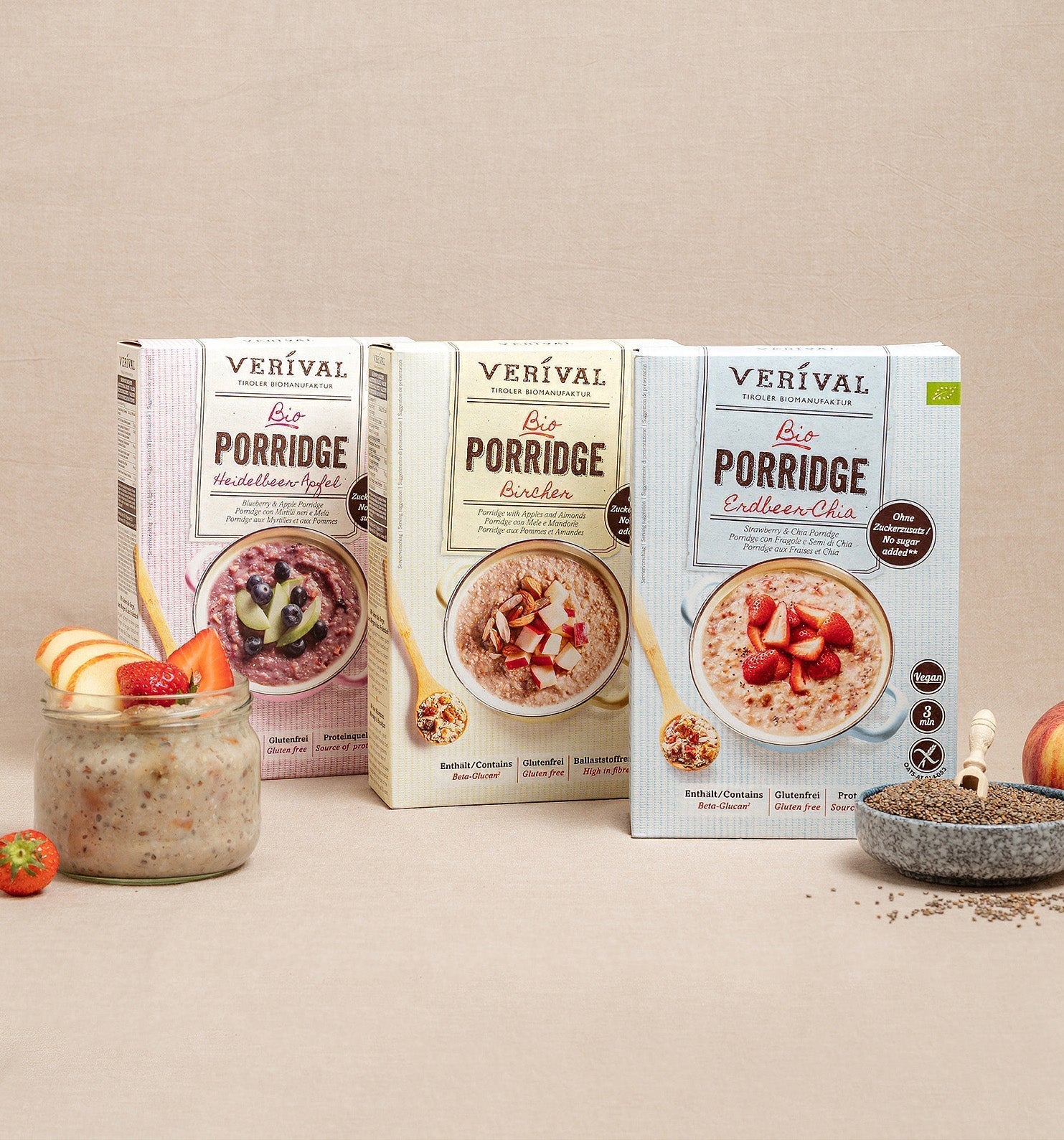 Drei Schachteln Verival Bio Porridge (Apfel-, Bircher- und Chia-Sorten) werden mit einem Glas Porridge, frischem Obst, Chia-Samen und einem Apfel auf einem beigen Hintergrund präsentiert.