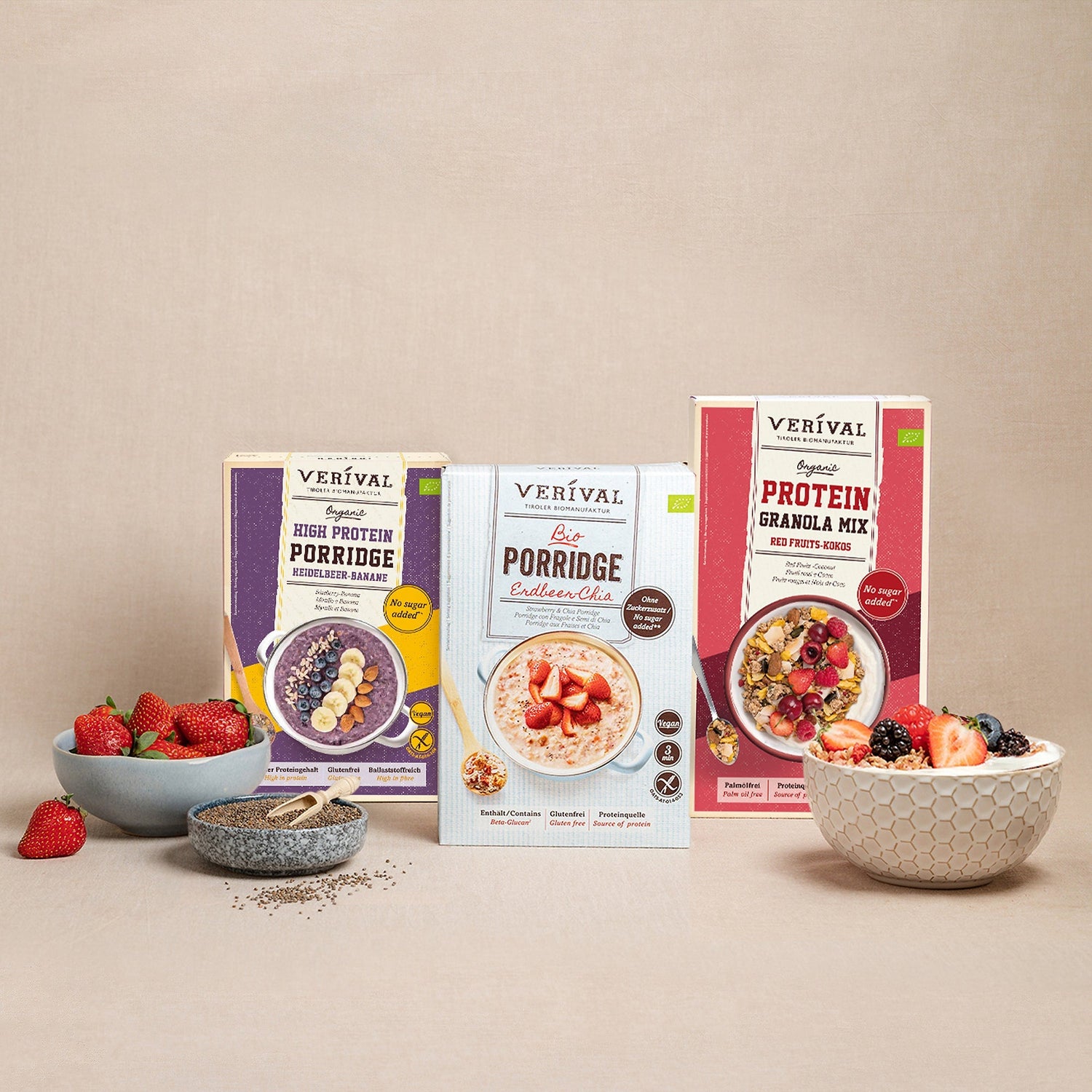 Ausgestellt werden drei Verival Porridge- und Granola-Boxen, umgeben von Schalen mit frischen Beeren und Chiasamen auf neutralem Hintergrund.