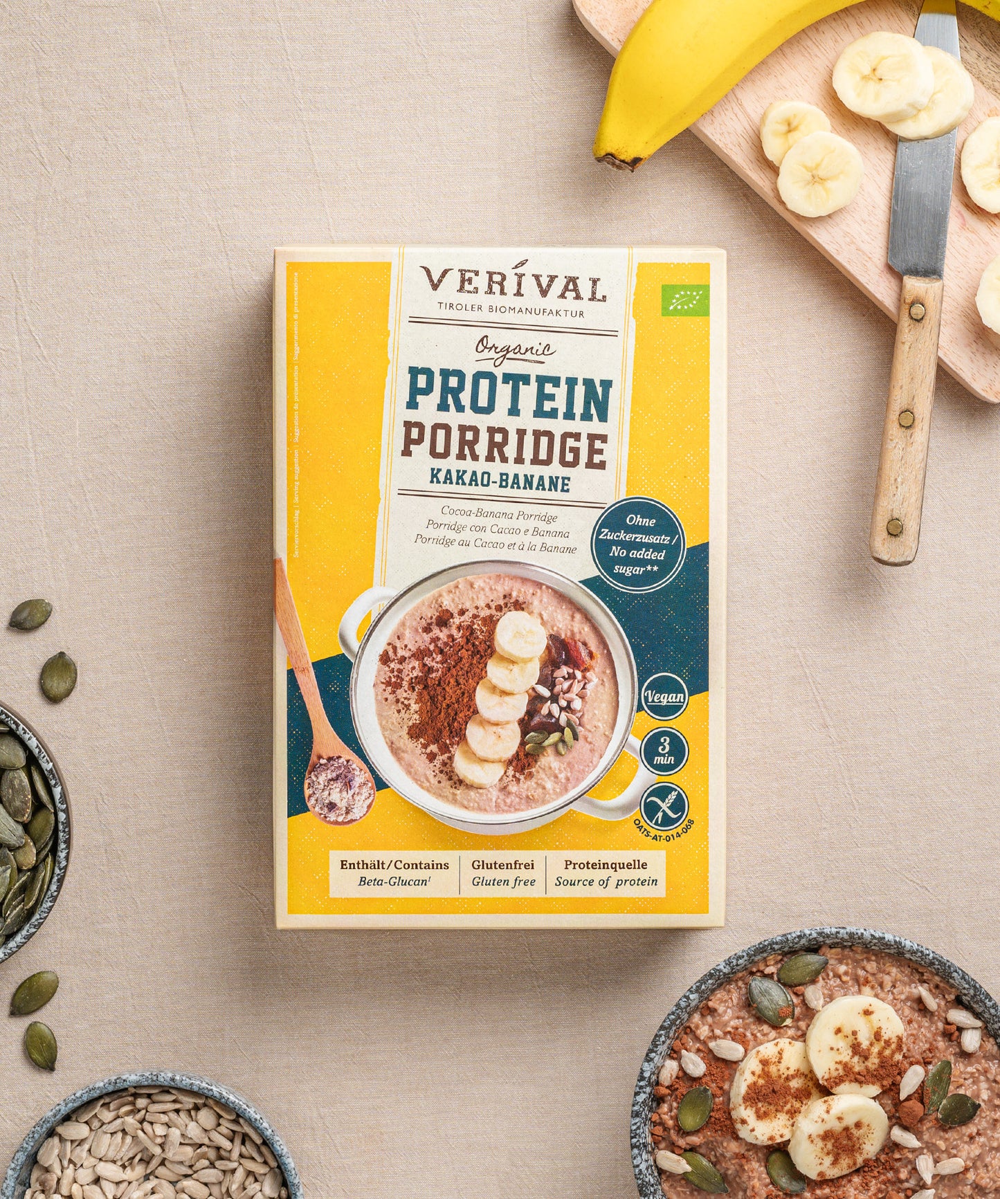 Eine Schachtel mit dem Set "Beste Frühstücksfreunde" der Verival Bio GmbH liegt auf einer beigen Fläche neben einer aufgeschnittenen Banane, einer Schüssel Porridge, Sonnenblumenkernen und Kürbiskernen.