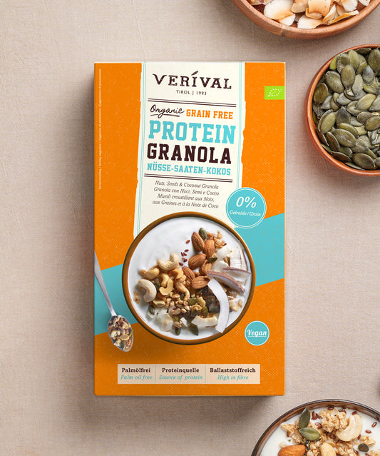 Eine Schachtel Bio Nuss-Kokos Grain Free Protein Granola der Verival Bio GmbH neben einer Schüssel Joghurt, garniert mit Nüssen, Samen und Kokosflocken, daneben zwei kleine Holzschälchen mit Kürbiskernen und Kokoschips.
