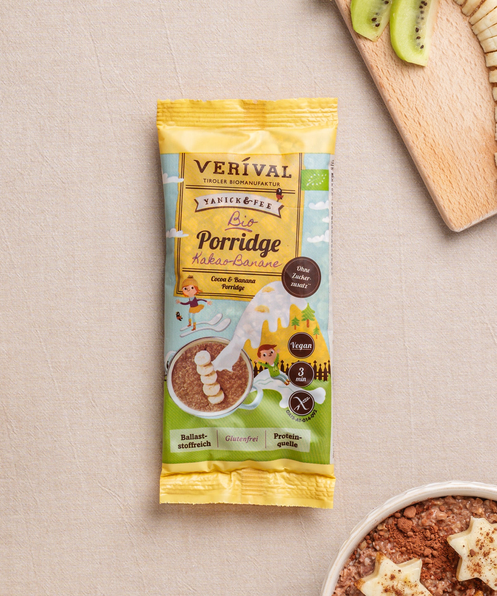 Eine Packung Kids Porridge Kakao-Banane von Verival Bio steht auf einem Tisch, dazu geschnittene Kiwis auf einem Holzbrett und eine Schüssel glutenfreies Porridge mit Bananenscheiben und sternförmigen Toppings, die Genuss und Ballaststoffe bieten.