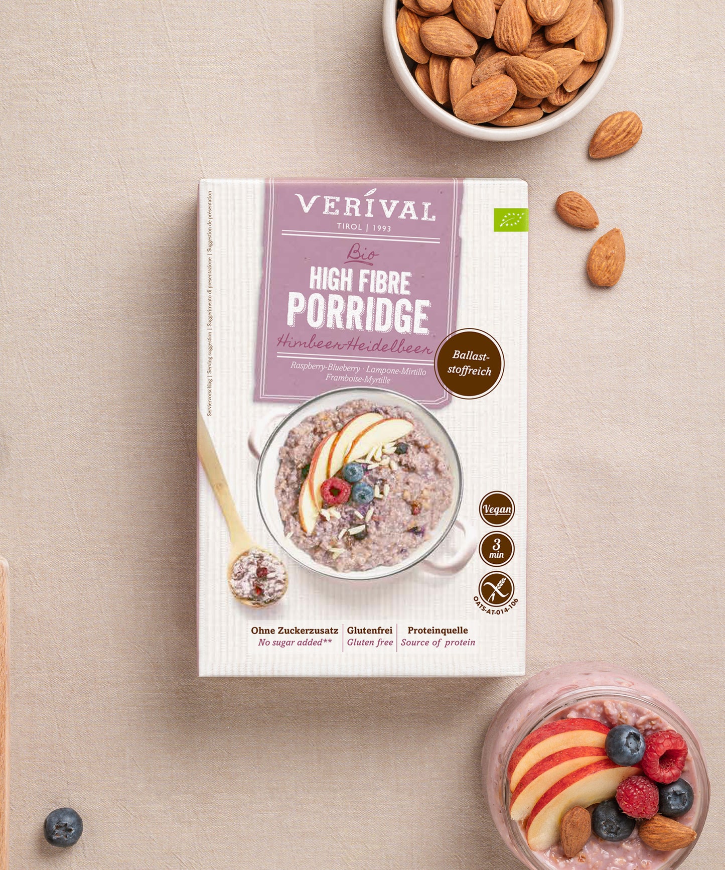 Eine Dose Bio High Fibre Porridge Himbeer-Heidelbeer der Verival Bio GmbH ruht auf einer beigen Fläche neben Mandeln und Früchtebrei - ein ideales veganes Frühstück voller präbiotischer Ballaststoffe.