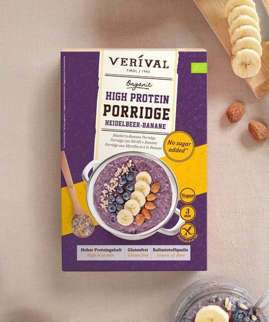 Eine Packung Verival Bio Bio Heidelbeere-Banane High Protein Porridge neben einer Schüssel mit Porridge aus violetten Beeren und Banane, umgeben von rohen Mandeln und Bananenscheiben.