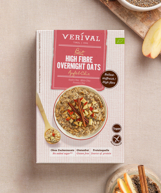 Eine Dose Verival Bio GmbH Bio High Fibre Oats Apfel-Chia steht auf einer beigen Unterlage mit Haferflocken, einer Apfelscheibe und einer Schale mit veganen Haferflocken und Chia - ideal für ein ballaststoffreiches Frühstück.