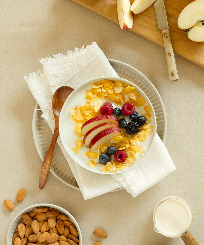 Eine Schüssel Joghurt mit Bio-Cornflakes (ungesüßt) von Verival Bio GmbH, Apfelscheiben, Himbeeren, Heidelbeeren und Brombeeren auf einer Serviette, dazu ein Holzlöffel. Daneben: Mandeln, Milchkännchen, Äpfel. Alles ohne Zuckerzusatz und glutenfrei.