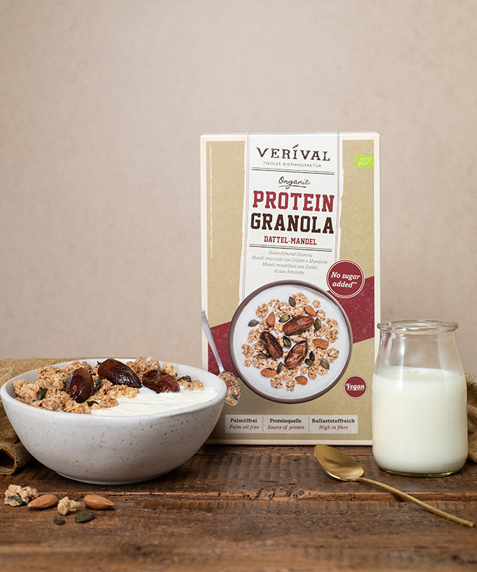 Eine Schüssel Bio-Dattel-Mandel-Protein-Müsli von Verival Bio GmbH, garniert mit Datteln und Mandeln, neben einer Packung des ballaststoffreichen Müslis und einem Glas Milch.