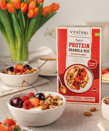 Vor einer Packung Bio Protein Granola Mix Red Fruits-Kokos der Verival Bio GmbH steht eine Schüssel Joghurt mit Kirschen, Erdbeeren, Müsli und roten Johannisbeeren. Im Hintergrund sind zwei weitere Schüsseln mit Müsli und Obst zu sehen.